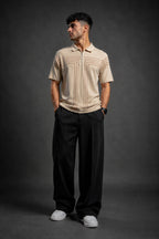A man modeling a beige short-sleeve knit polo shirt with vertical stripe details, paired with black trousers and white sneakers, standing against a dark gray studio background.