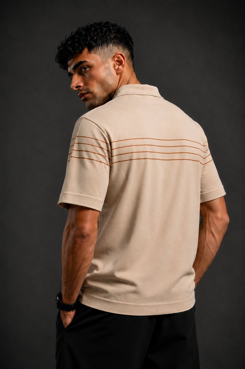 A man wearing a beige short-sleeve polo shirt with thin horizontal brown stripes across the upper back, shown from a three-quarters back view against a dark gray studio background.