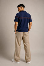 A man is shown from the back wearing a navy blue short-sleeve polo shirt with thin horizontal red stripes across the upper back and shoulders, paired with beige trousers and white sneakers, against a solid light gray studio background.