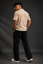 A man wearing a beige short-sleeve polo shirt with thin horizontal stripes on the chest, paired with black trousers and white sneakers, standing against a dark gray studio background.