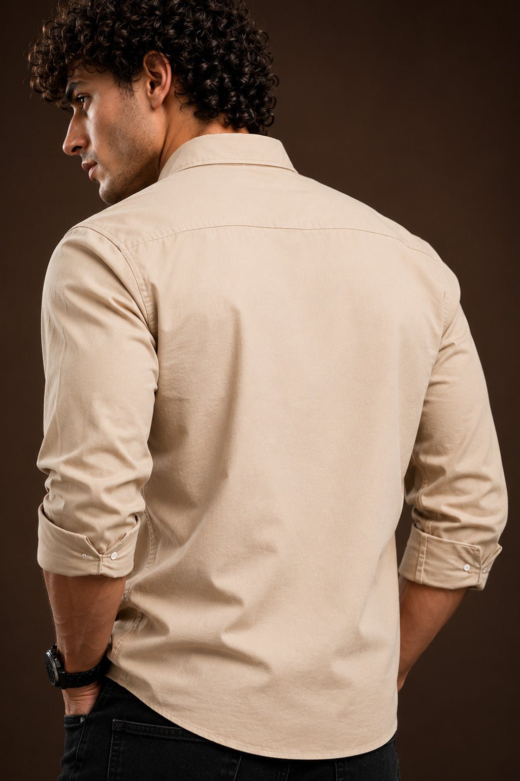 A man wearing a light beige long-sleeve button-down shirt, shown from the back against a dark brown studio background.