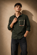 A man wearing a dark olive green long-sleeve button-up shirt with a multicolor plaid patterned collar and chest pocket, paired with dark jeans, standing against a textured beige wall.