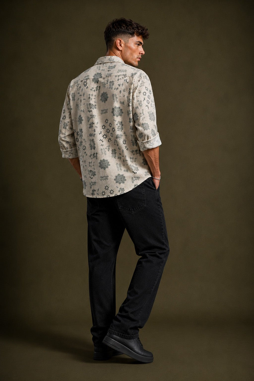 A man is shown from the back wearing a light beige long-sleeve button-down shirt with a dark gray floral pattern, paired with black trousers and black shoes, set against a dark brown studio background.