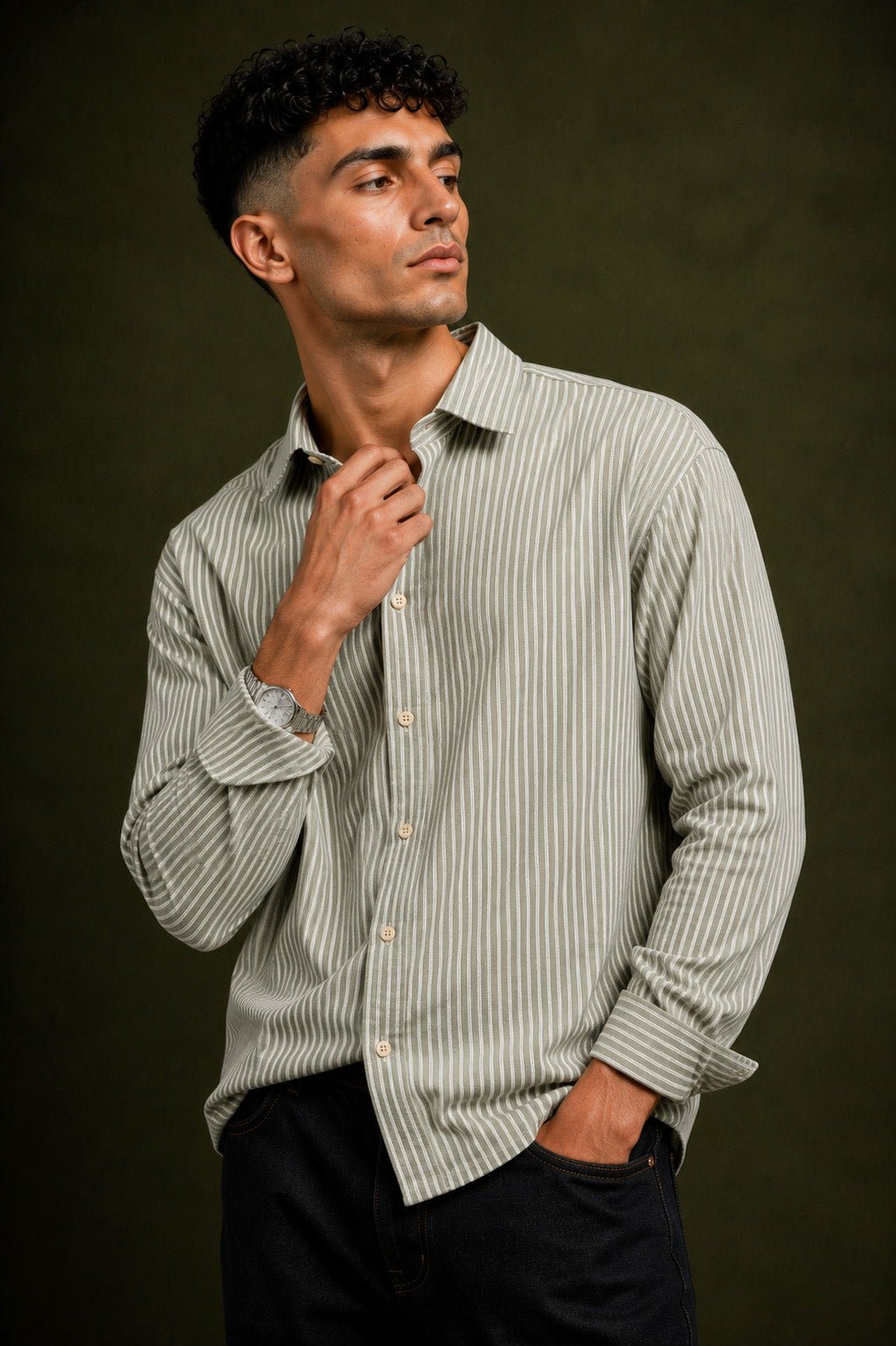 A man wearing a light beige and dark green vertically striped long-sleeve button-down shirt, paired with black trousers, posed against a dark olive green background.