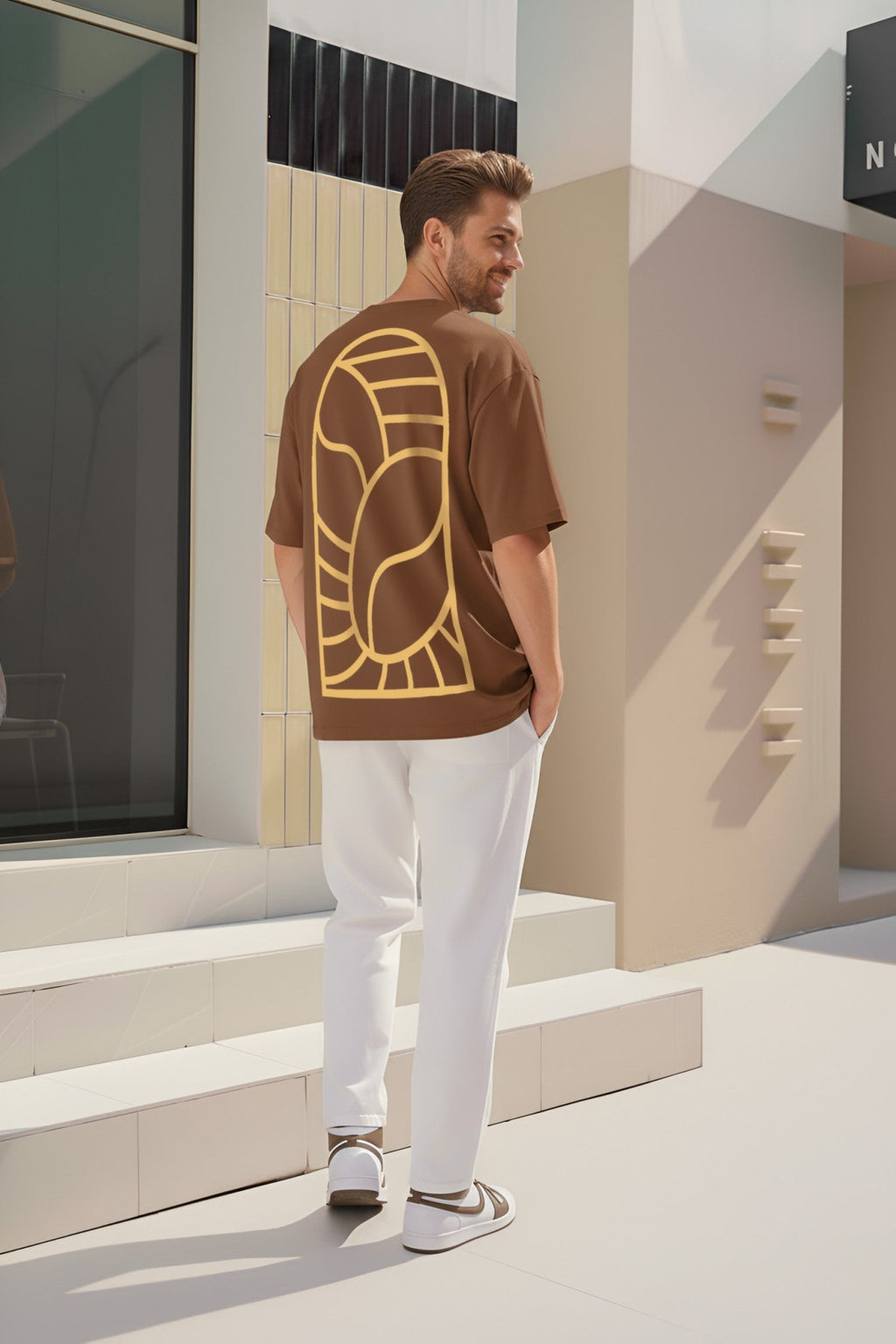 Back view of Mocha Maniac brown oversized T-shirt featuring minimal golden abstract back print, worn by male model in outdoor setting