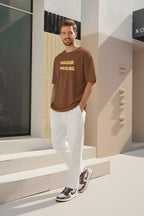 Front view of Mocha Maniac brown oversized T-shirt with bold yellow text logo, styled by male model with white pants and brown sneakers