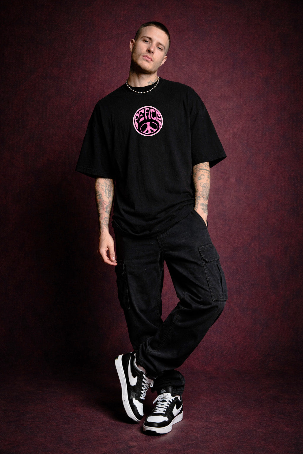 Man wearing a black t-shirt with a pink logo and black pants against a dark red background
