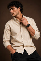 A man wearing a light beige long-sleeve button-down shirt and black pants, posing against a dark brown studio background.