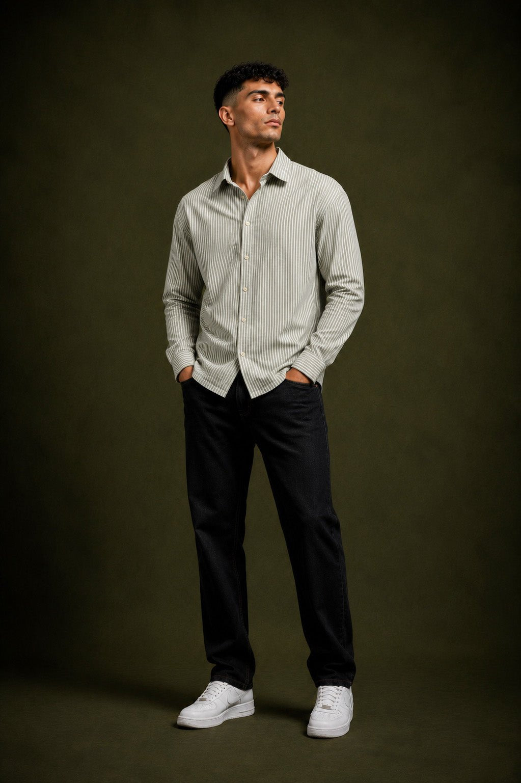 A man wearing a light gray and white vertically striped long-sleeve button-down shirt, black trousers, and white sneakers, standing against a dark olive green studio background.