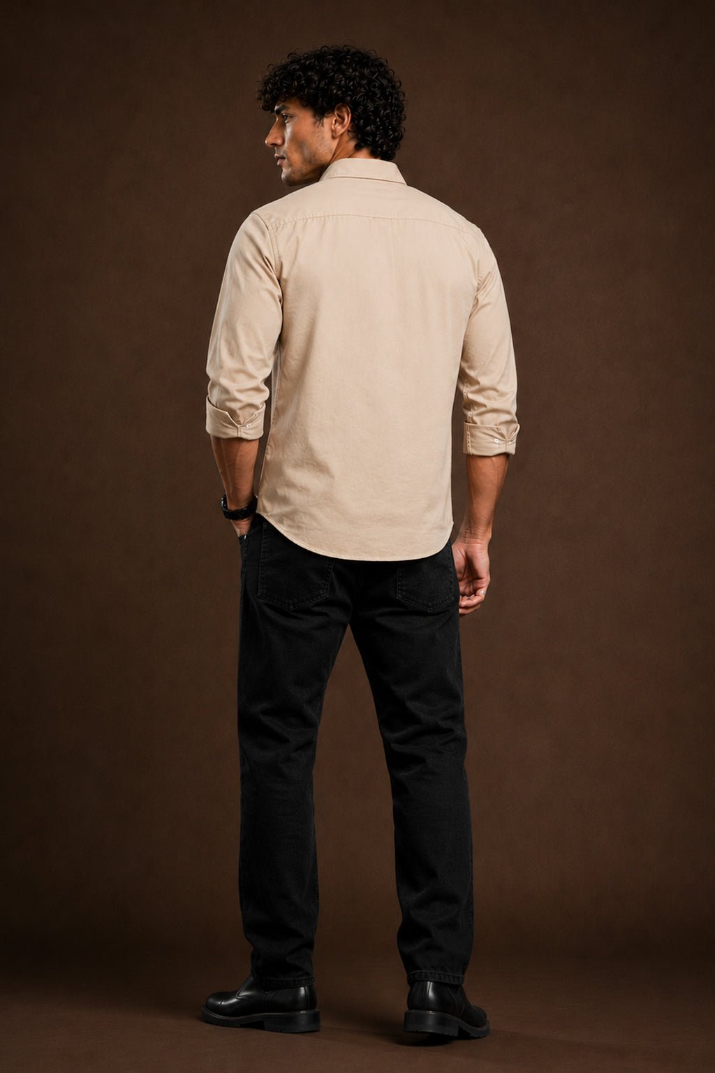 A man shown from the back wearing a light beige long-sleeve button-down shirt, black trousers, and black shoes, standing against a solid dark brown studio backdrop.