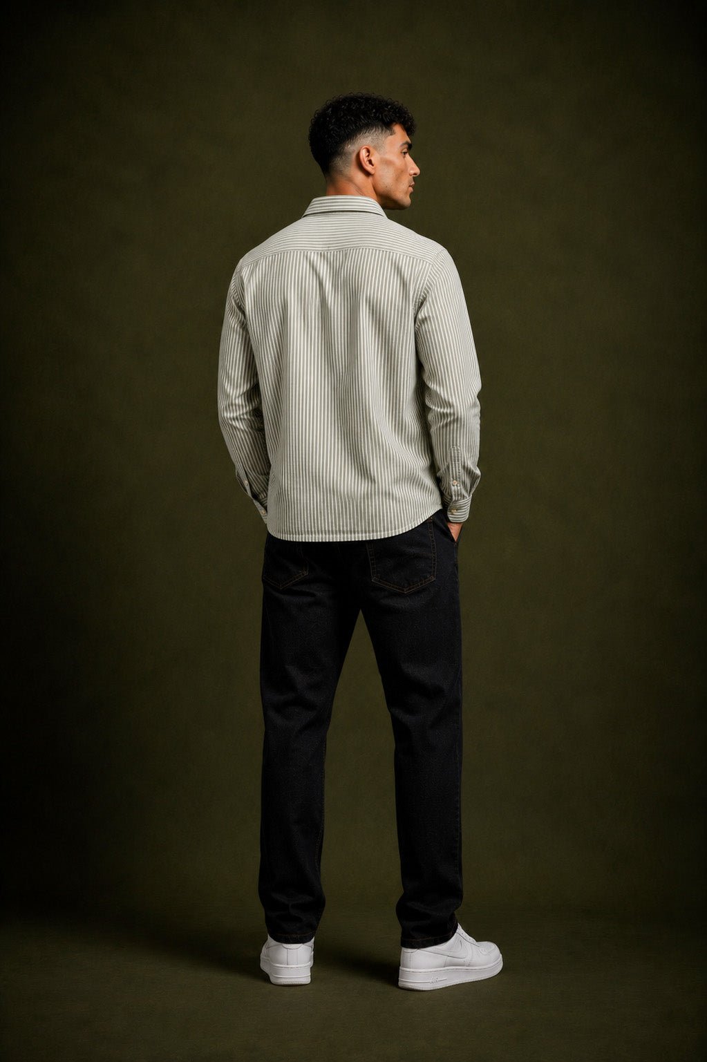 A man standing with his back to the camera, wearing a light beige and white vertically striped long-sleeve button-down shirt, black trousers, and white sneakers, set against a dark olive green studio background.