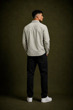 A man standing with his back to the camera, wearing a light beige and white vertically striped long-sleeve button-down shirt, black trousers, and white sneakers, set against a dark olive green studio background.